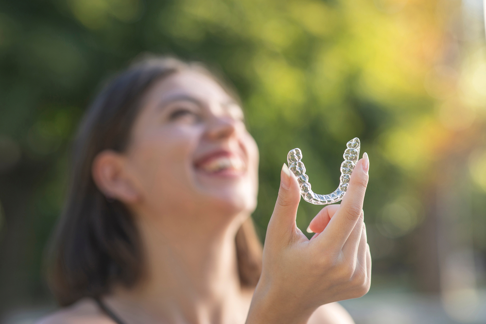 Young woman smiling while holding clear aligners in natural sunlight, highlighting the discreet and modern orthodontic treatment available with Invisalign in Cupertino - Invisalign Cupertino