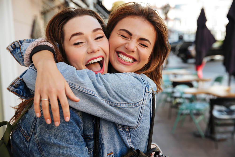 Two young women sharing a joyful moment and showing radiant smiles, representing the confidence and natural beauty achieved with porcelain veneers in Cupertino - Porcelain Veneers in Cupertino