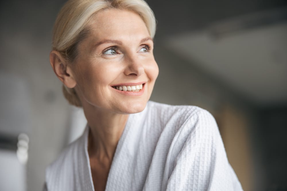 A smiling woman with short blonde hair, wearing a white textured top, gazes off to the side in a softly lit environment - dentist Cupertino