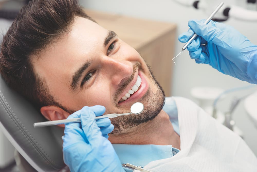 A smiling man reclines in a dental chair while a dentist, wearing blue gloves, holds dental tools near his mouth - dentist Cupertino
