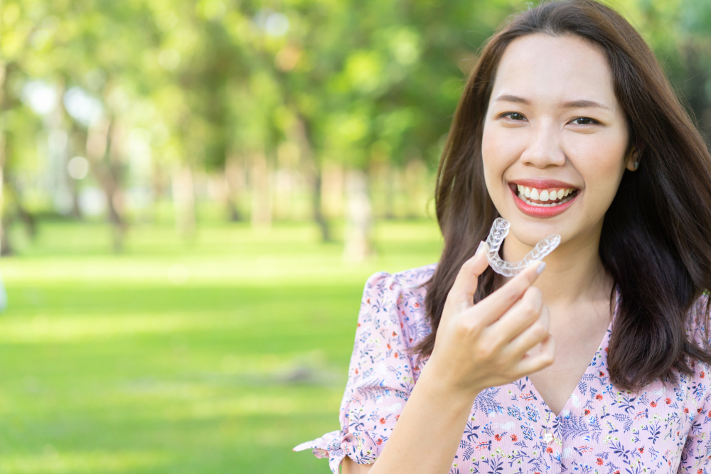 Portrait of a smiling woman holding transparent aligners in a park setting, representing comfortable and nearly invisible orthodontic treatment with Invisalign in Cupertino - Invisalign Cupertino