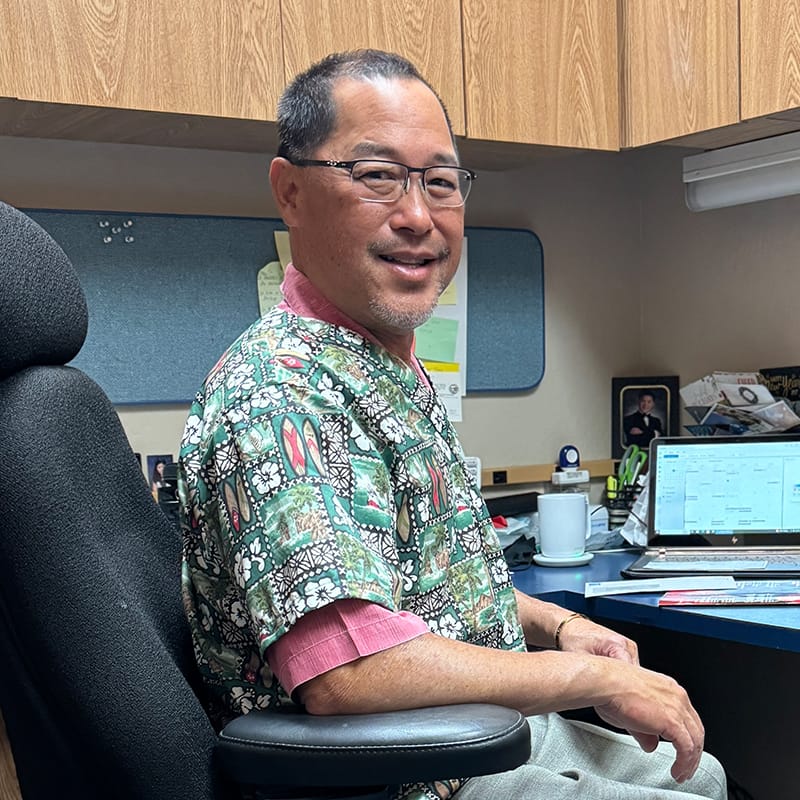 Dr. Jeffrey K. H. Ching, DDS, is seated in his office, wearing glasses and a patterned scrub top, with a computer and desk in the background - dentist Cupertino