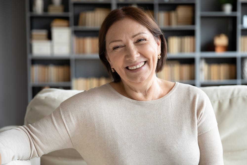 A cheerful older woman sitting on a sofa, smiling warmly, with a bookshelf filled with books in the background - dentist Cupertino