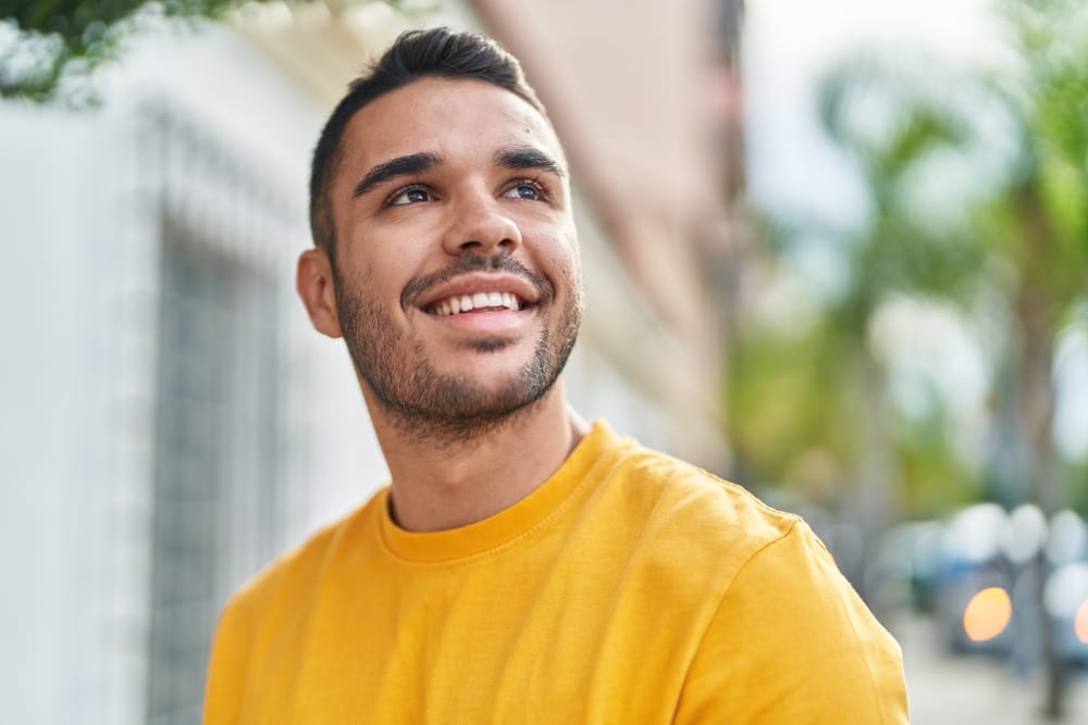 A smiling young man in a bright yellow shirt looking up and to the side, standing outdoors with a blurred urban background featuring greenery and buildings - dentist Cupertino