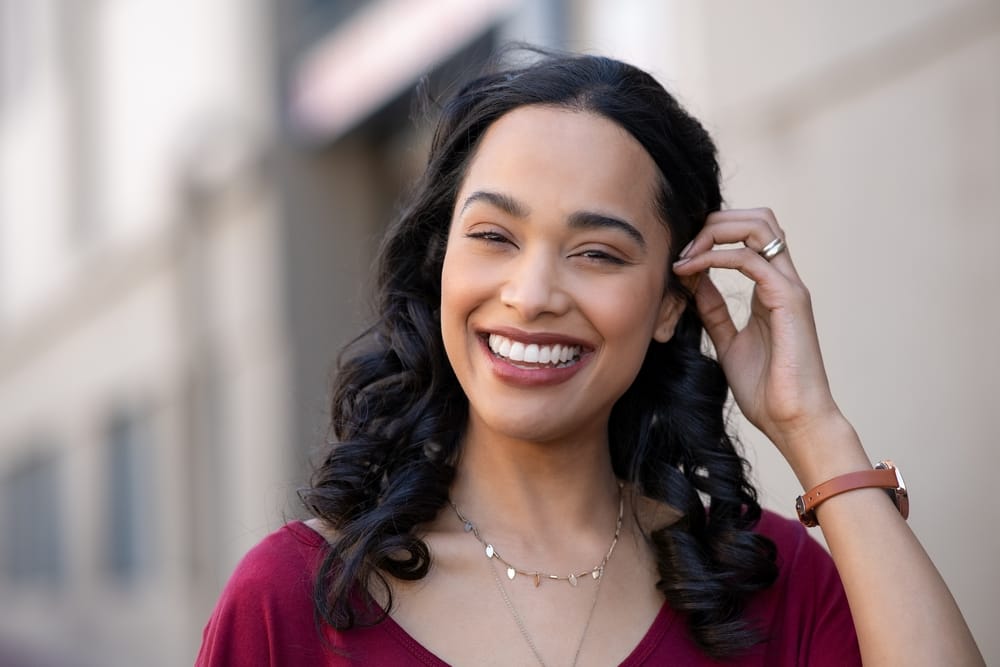 A cheerful woman with dark, wavy hair, wearing a red top and delicate jewelry, smiling brightly outdoors with a blurred urban backdrop - dentist Cupertino