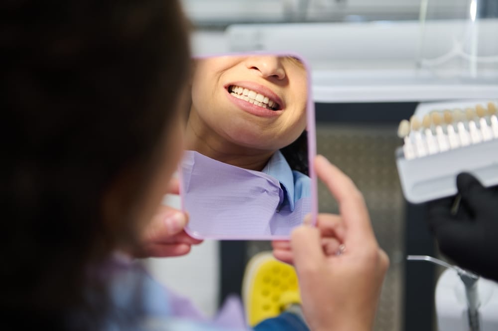 A person examines their smile in a handheld mirror during a dental visit, with a dentist holding a shade guide in the background - dentist Cupertino